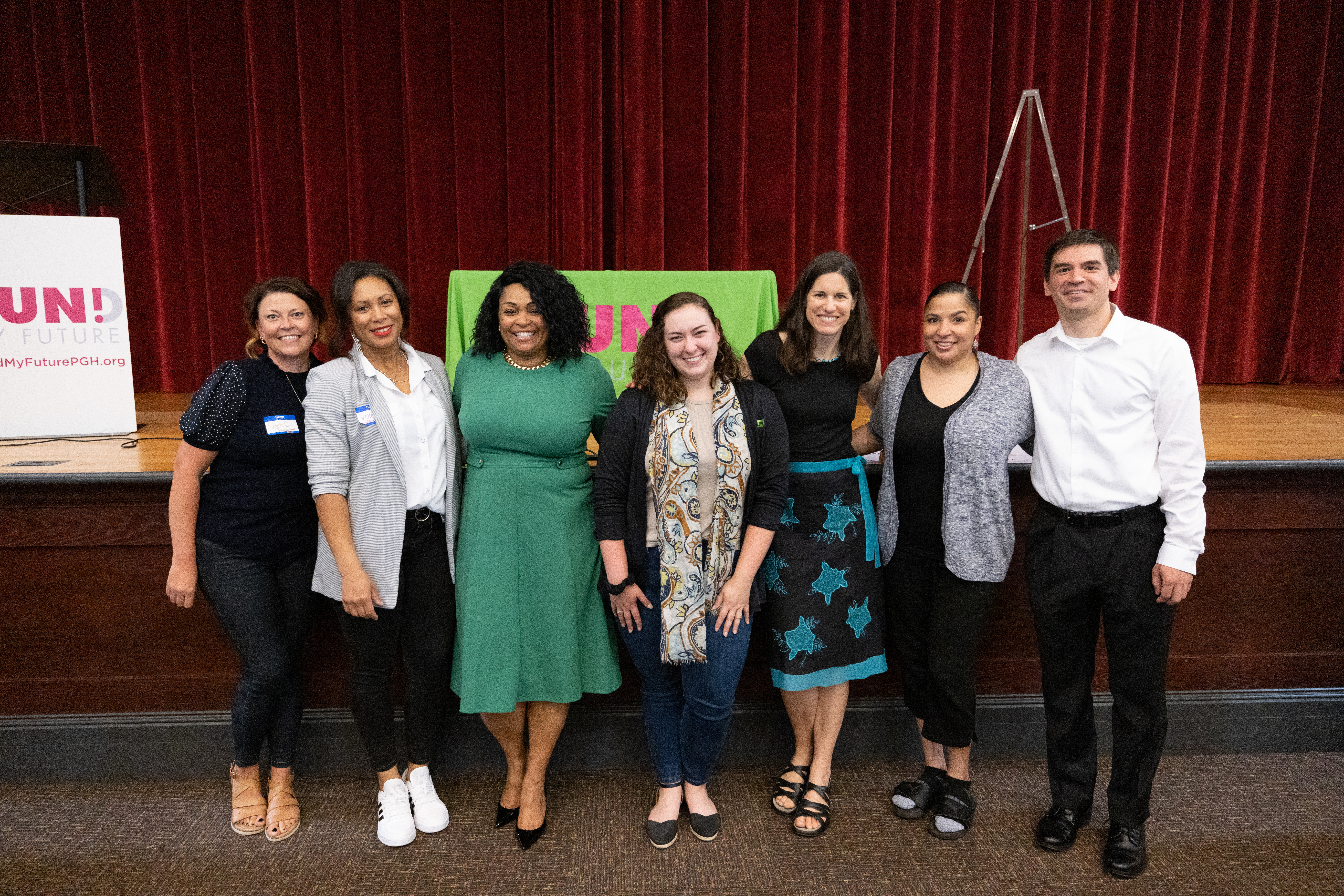 7 Neighborhood Allies staff members - 6 women and 1 man - gather for a posed photo in front of the stage at our annual Fund My Future PGH celebration.