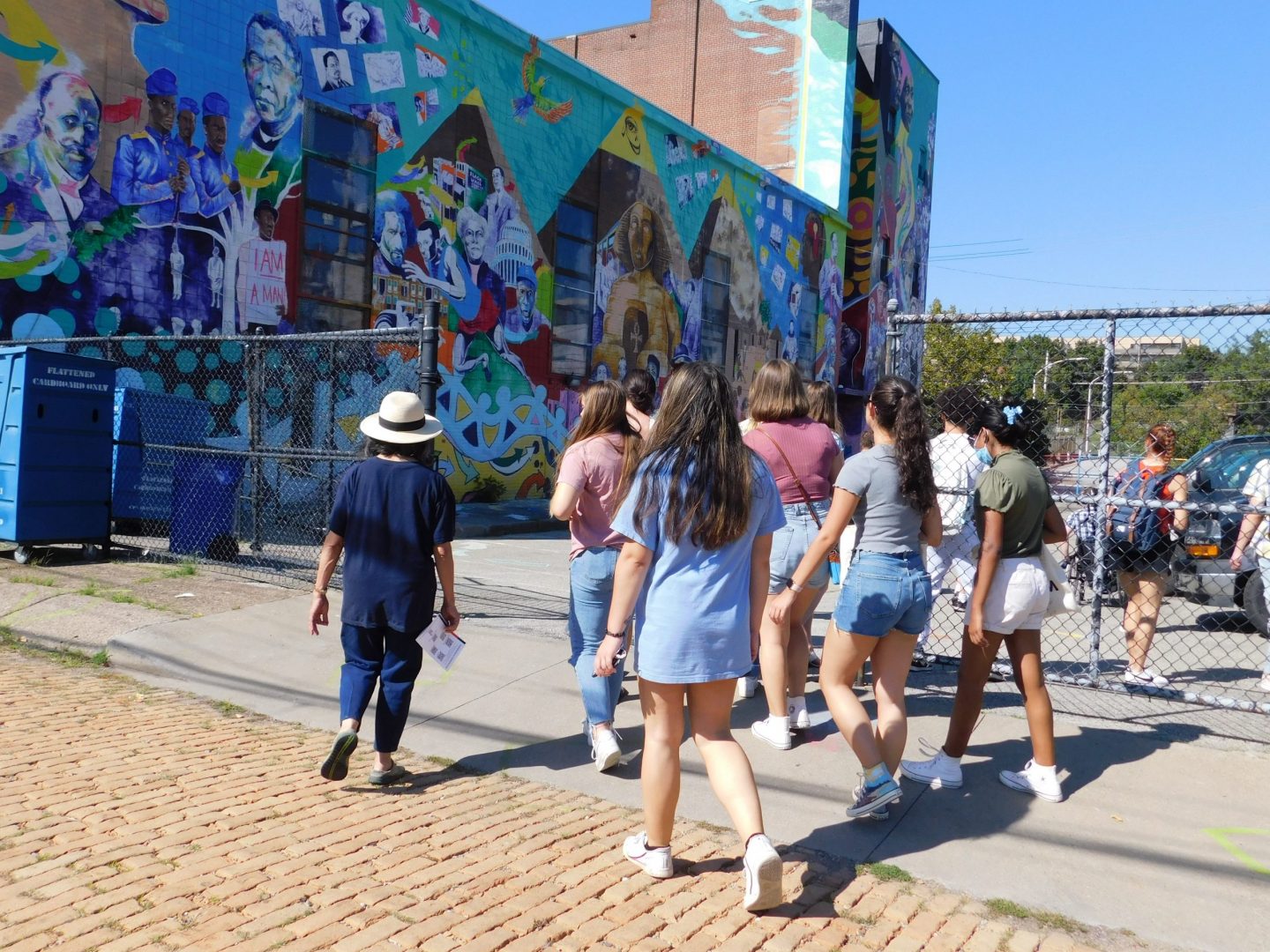 Image of a group of people walking into the current parking lot (future site of the CEA plaza). The people are walking towards the colorful mural that takes up the entire side of the CEA building.