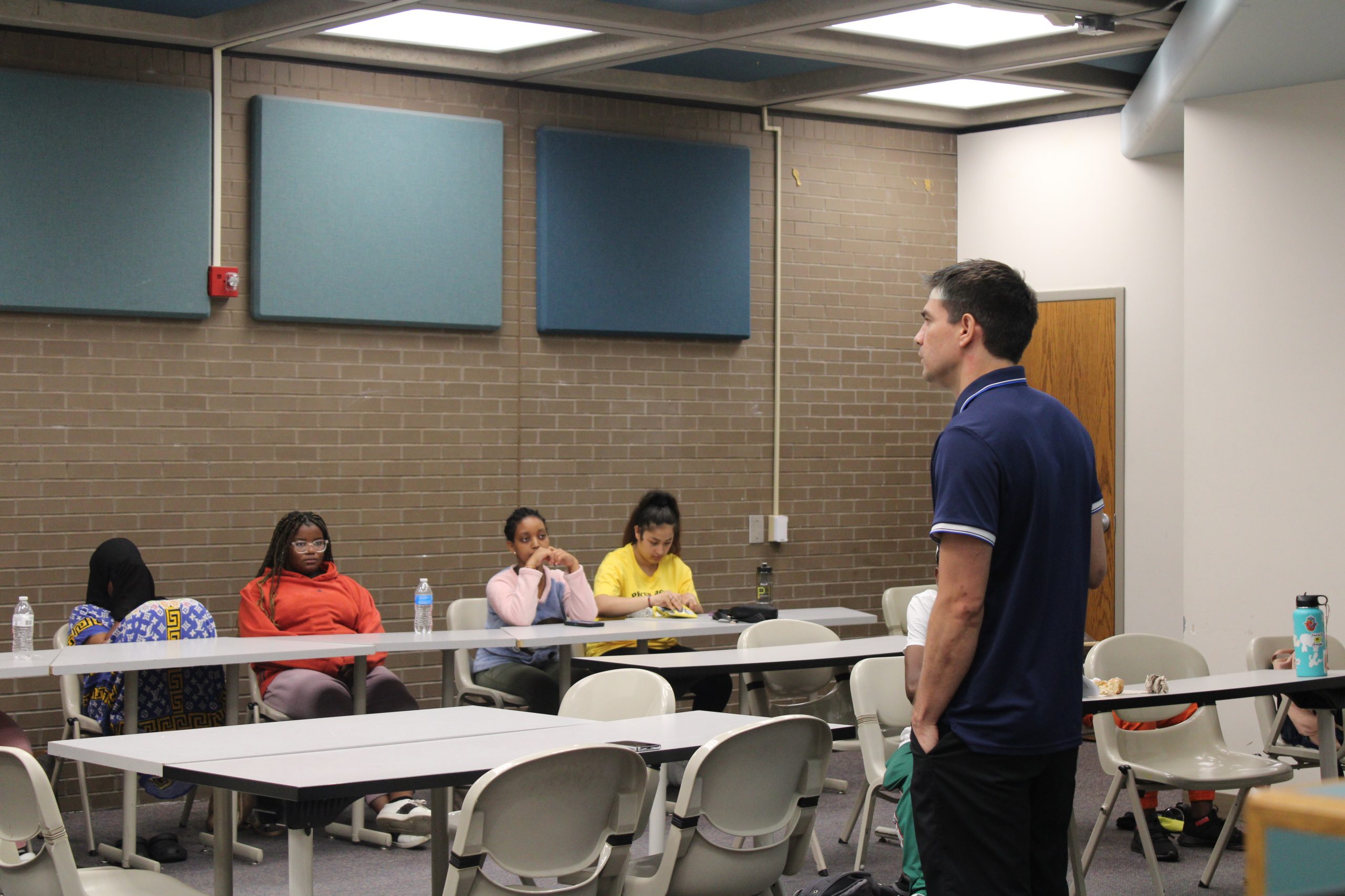 Image of Javier, our staff person, teaching a class of teens about financial education. A row of teens sit along a long grey table at the back of the room, while we see Javier's back. He wears a blue polo shirt.
