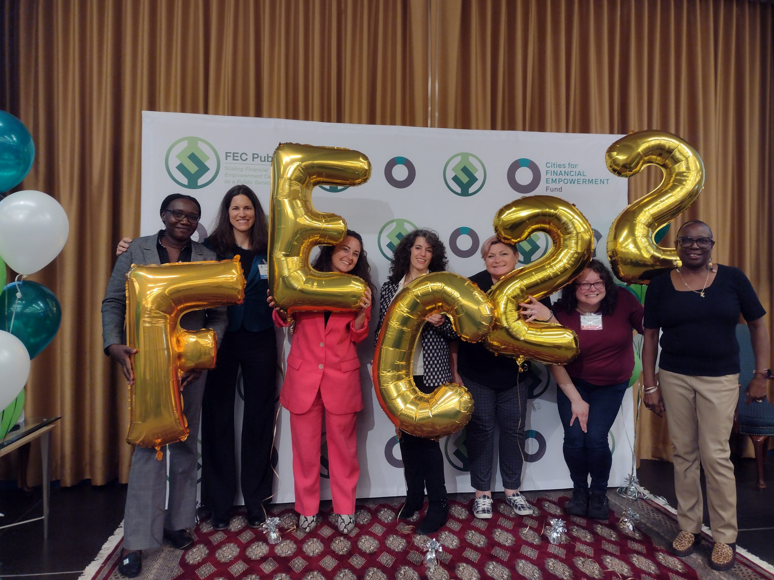 Image of our 5 Financial Empowerment Center (FEC) counselors and the Director of the FEC (all are women) with Sarah, our Director of Economic Opportunity. They all stand in front of a backdrop with the green FEC logo. They hold gold, shiny balloons that spell out "FEC 22." They are all smiling.