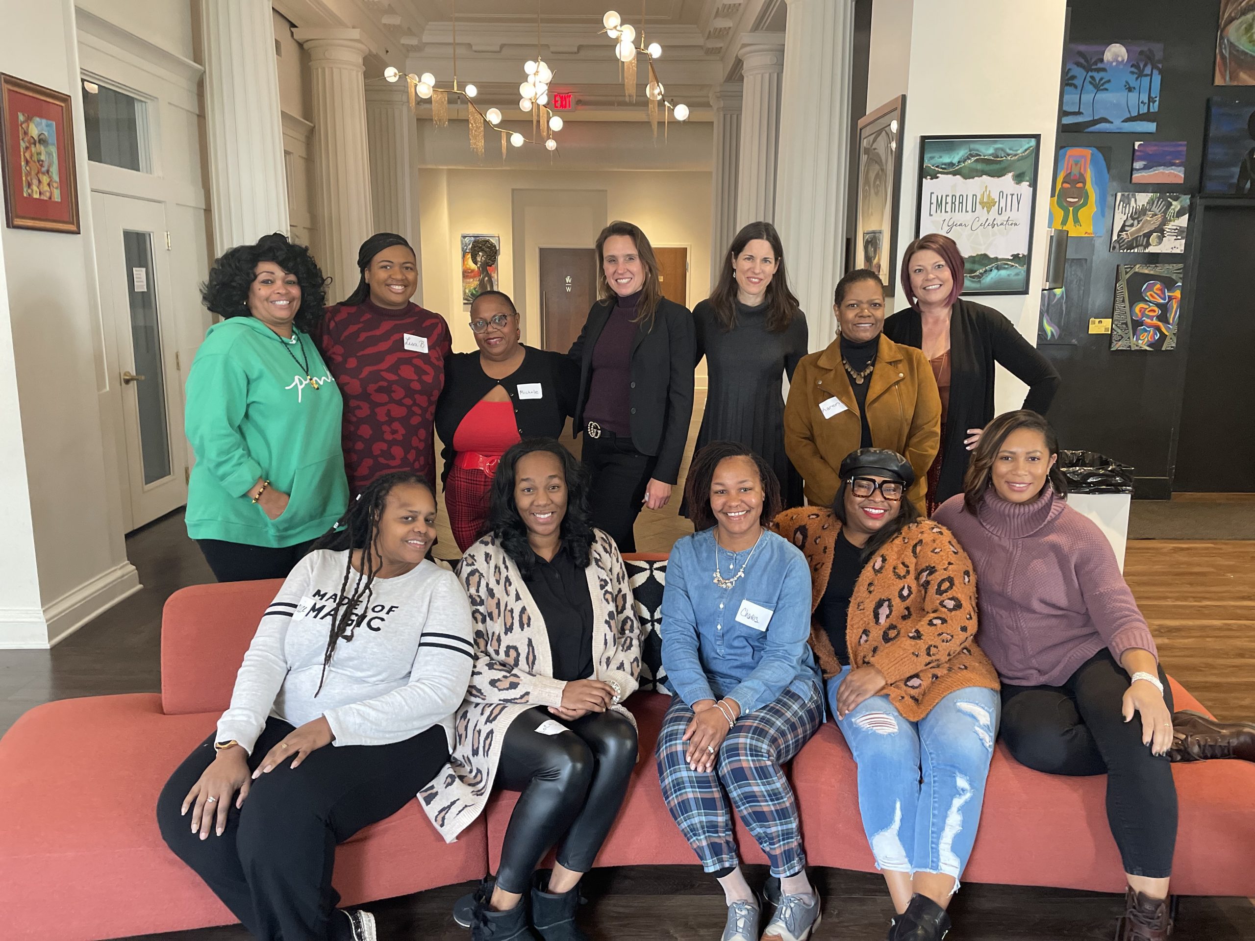 Image of participants, instructors, and Neighborhood Allies staff of the Money Talks program. 5 Black women sit on a light red couch, while 7 more women - 5 Black and 3 white - stand behind the couch. They all smile and pose for the camera. Behind them is a large room with tall ceilings, a chandelier, and white pillars.