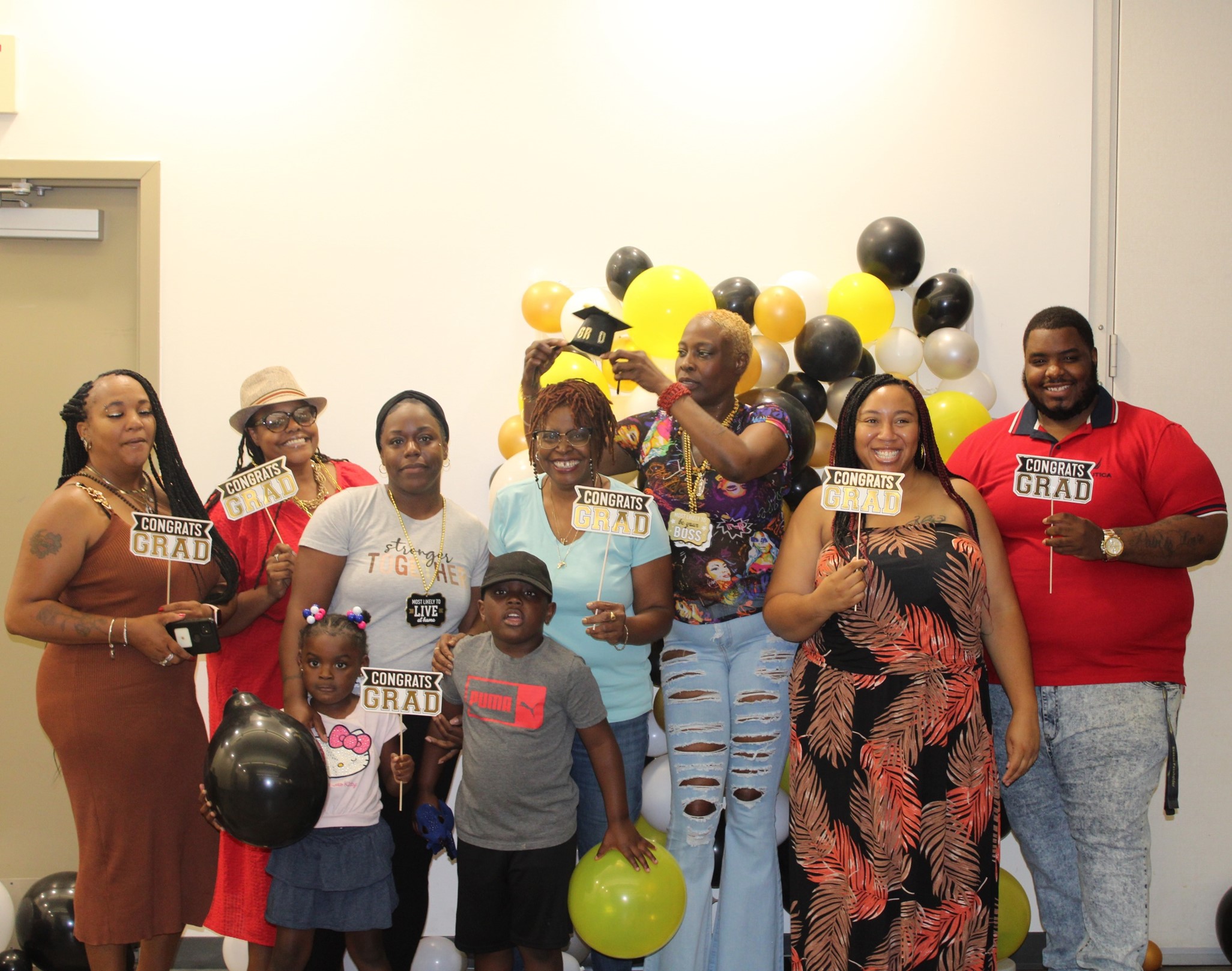 Image of Beams to Bridges participants (one of Steel Smiling's programs) at their graduation. Everyone stands smiling at the camera, some holding photobooth props such as a mini graduation cap, balloons, or signs that say "congrats, grad!" Behind them is a short balloon arch made of black and yellow ballons.