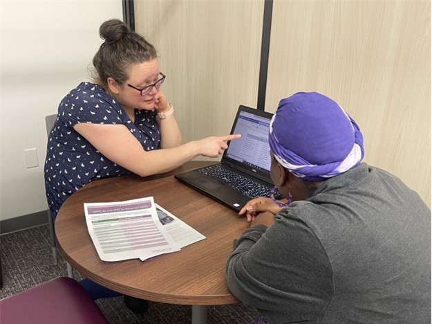 Image of Alicia - a financial counselor and white woman wearing a blue polka dot short sleeve blouse and glasses - pointing at a computer screen beside her. A black woman wearing a purple head wrap and grey sweater, looks on. The pair sit at a circular table.