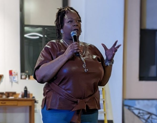 Image of Steel Smiling staff member, Robin Sheffey, a Black woman, wearing a brown faux leather short sleeve shirt and jeans, her dark hair up in a ponytail. She holds a microphone and gestures with her left hand while giving a speech.